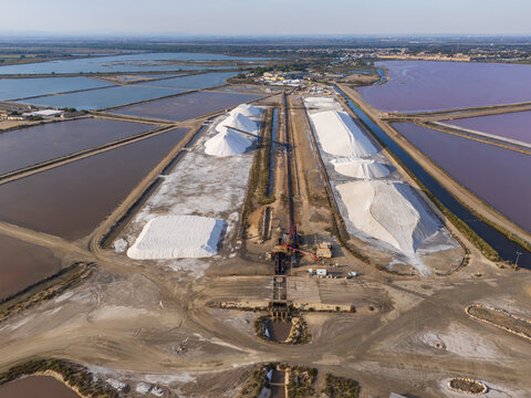 Aerial view of the sea salt works featuring large white salt piles and evaporation ponds with pink water in Aigues-Mortes, Occitanie, France.