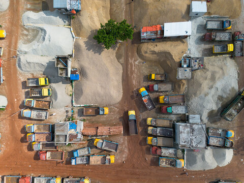 Aerial view of a busy open-air brick and sand market with numerous trucks parked among large piles of construction materials and a single tree in Dhaka, Dhaka, Bangladesh.