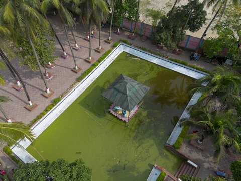 Aerial view of the Sri Sri Loknath Baba Mondir featuring a central shrine structure set within a green pond, surrounded by palm trees and paved walkways in Narayanganj, Dhaka, Bangladesh.