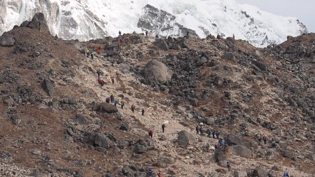 Large group of trekkers and mountaineers with backpacks slowly ascending a steep, rocky trail against of majestic snow-capped himalayan peaks during an expedition. Everest base camp trek in Nepal