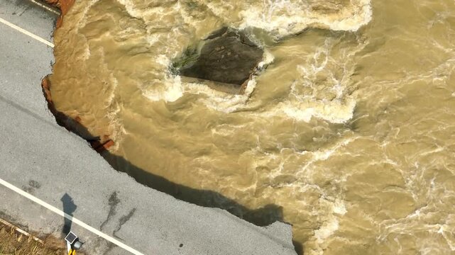 Aerial view captures a severely damaged road washed away by flash flooding, exposing eroded soil and broken asphalt as powerful muddy water destroys transportation infrastructure. Thailand. 4k.
