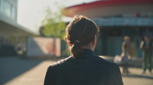 Woman sits on a bench facing an urban plaza as warm late afternoon sunlight casts long shadows, passersby and storefronts create a city scene with contemplative, solitary mood. 4K.