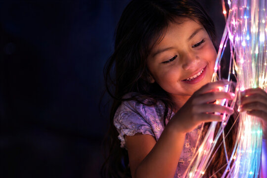 Smiling hispanic little girl touching glowing fiber optic light strands in dark sensory room, face lit with colorful light, wonder