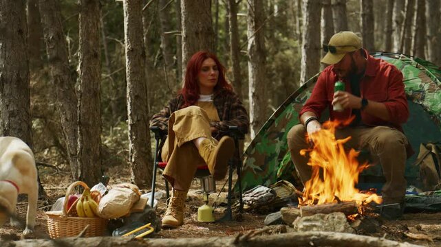 Couple relaxes by a campfire while camping in a pine forest, drinking from a can and mug beside their tent and dog.