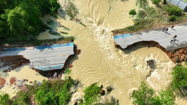 Aerial photography reveals a roadway completely severed by floodwater, exposing damaged asphalt and washed away ground, emphasizing soil erosion, disaster recovery, and climate resilience. Thailand.
