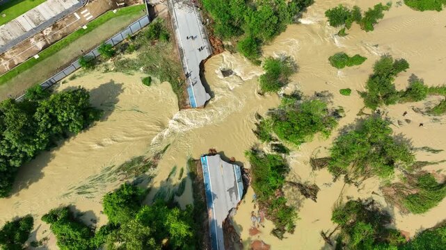Aerial view captures a severely damaged road split apart by powerful floodwaters with muddy currents rushing through the collapsed section, highlighting natural disaster impacts and road safety risks.