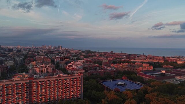 Warm sunset light bathes dense Eixample rooftops stretching toward Mediterranean coastline, modernist hospital dome and distant towers framing Barcelona city skyline. Aerial drone wide shot