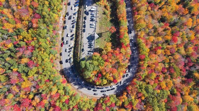 Tourist cars line hairpin curve on Kancamagus Highway, circling through blazing orange autumn foliage within dense White Mountain National Forest hillside, New Hampshire. Aerial drone top down shot