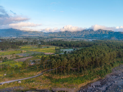 Drone panorama of agricultural fields, rural homes, forested mountains, drifting clouds, and a winding road near Pronojiwo, Indonesia.