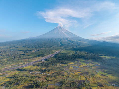 Drone panorama of patterned agricultural fields beneath Mount Semeru, with forested slopes, volcanic terrain, and expansive blue sky.