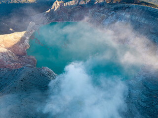 Clouds drift over a turquoise volcanic crater lake. Rugged, gray slopes encircle the glowing water at Mount Ijen © Cloudyew