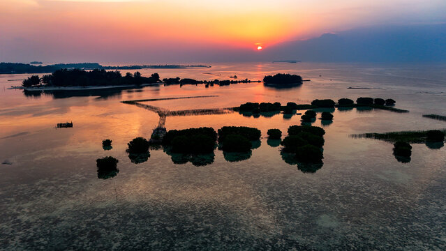 Aerial view of Pari Island at sunset, featuring calm waters, small mangrove clusters, and a vibrant orange sky over the Thousand Islands in South Kepulauan Seribu, Jakarta, Indonesia.