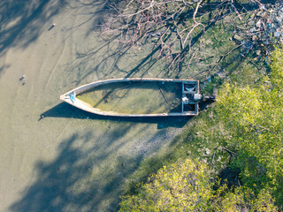 Lonely boat rests on calm, murky water. Branches cast shadows across the surface. Green foliage frames the scene's edge © Cloudyew
