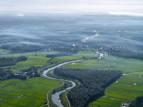 Misty morning blankets the green valley below. River winds through fields, cutting a graceful path at Kubang Semang
