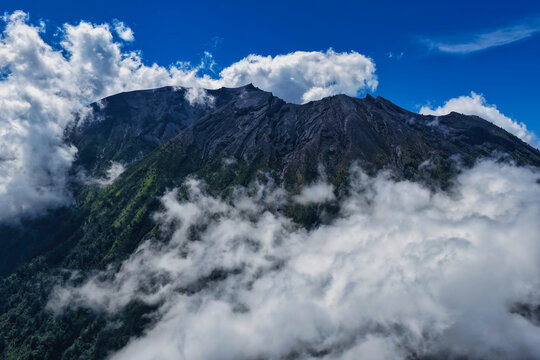 Aerial view of Mount Agung with its rugged volcanic slopes partially covered by clouds under a clear blue sky in Bebandem, Bali, Indonesia.