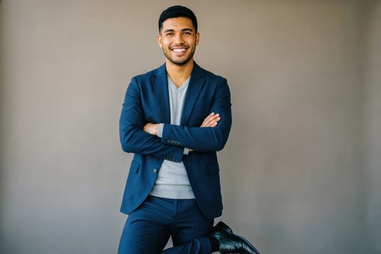 Un joven hispano est&aacute; de pie frente a una pared lisa de color gris c&aacute;lido, sonriendo abiertamente y con los brazos cruzados. Luce seguro y esperanzado mientras piensa en sus metas futuras