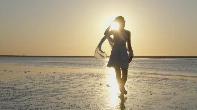 Backlit woman in white dress walking barefoot across wet sand