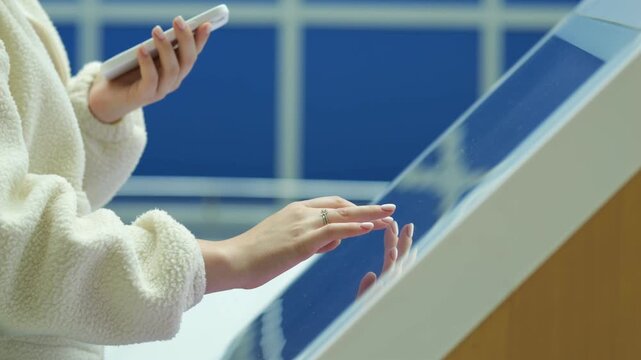 Person holds phone while touching large interactive screen. Hands move across glass surface. Background shows blue tiled wall and wooden structure. Person wears soft textured jacket
