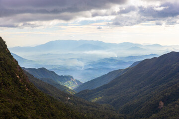 Layered mountain ridges descend into a mist-filled valley framed by dense forest slopes and a dramatic cloudy sky, evoking calm, remoteness, and natural beauty for travel and nature themes. Vietnam © evannovostro