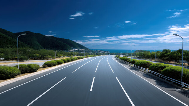 Wide two way roadway with clear lane markings, surrounded by green trees, blue sky, and distant mountains