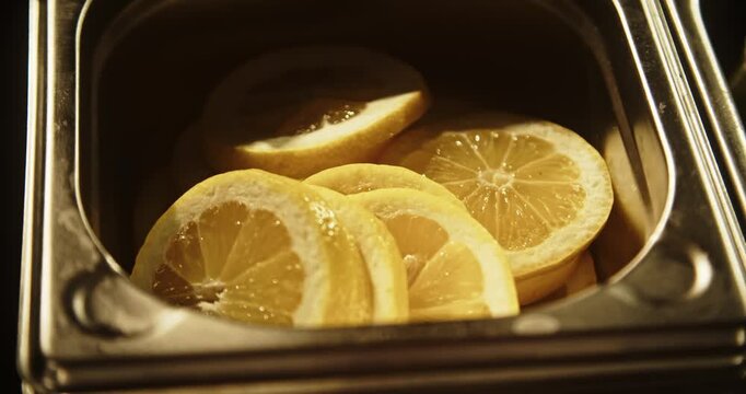 Close up of fresh juicy lemon slices arranged in a stainless steel container at a cocktail bar. Warm golden lighting highlights the citrus texture and professional drink preparation setup.