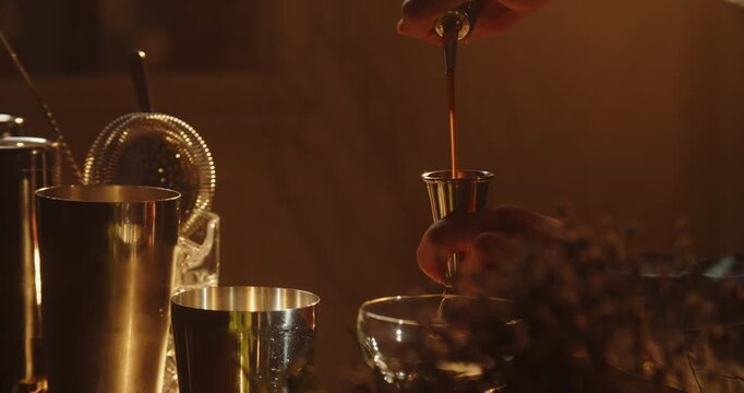 Close up of a bartender carefully pouring a liquid ingredient into a metal jigger. Warm ambient lighting and blurred bar equipment create an intimate cocktail preparation scene.