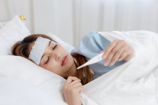 Sick young woman resting in bed checking fever with thermometer feeling exhausted and unwell with cooling patch for cold symptom