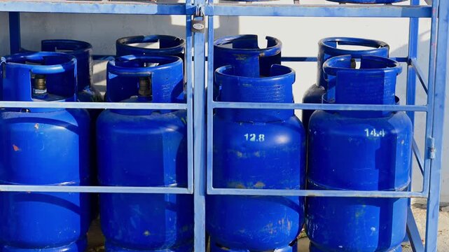 A row of blue liquefied petroleum gas cylinders is stored securely inside a locked metal cage under bright sunlight.
