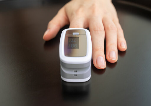 Finger inserted into pulse oximeter, oxygen saturation 86 and heart rate 77 on screen. Fingertip sensor measures SpO2 and vital signs on dark glossy surface. Portable medical gadget for home