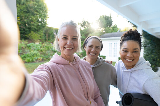 Female friends posing for selfie on porch, wearing hoodies, holding smartphone, rolled yoga mat