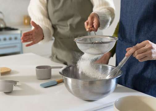 Two women wearing aprons sifting flour through mesh sieve into metal bowl in kitchen, whisking