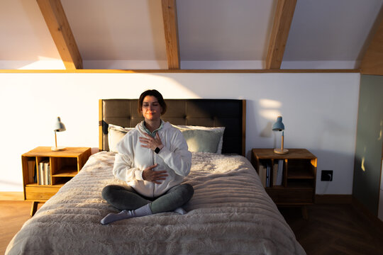 Woman sitting cross-legged on bed in bedroom wearing zip-up hoodie and leggings, showing smartwatch