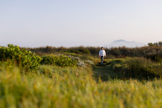 Woman walking through tall grasses along narrow path in dunes, wearing white top, dark pants