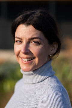 Woman smiling, facing camera, wearing light gray ribbed turtleneck sweater gold hoops in garden