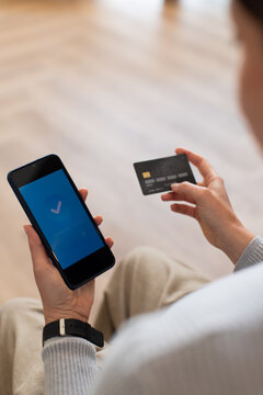 Female sitting at home holding smartphone showing blue checkmark and EMV card wearing watch