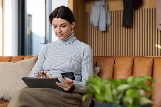 Female wearing light gray turtleneck sitting on tan leather sofa holding tablet and checking card