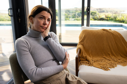 Woman sitting in armchair in living room, wearing grey turtleneck, facing sliding glass doors