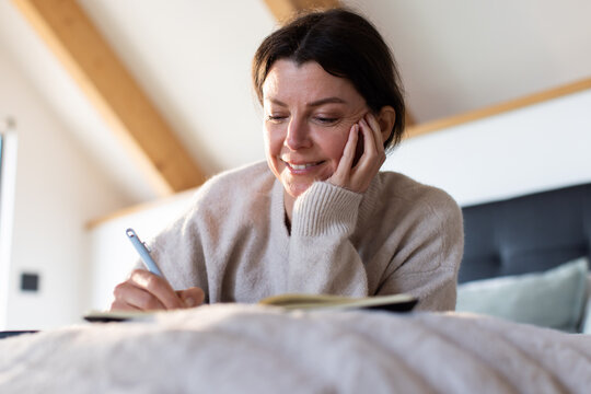 Woman reclining on bed writing in open notebook with pen in loft bedroom, wearing beige sweater