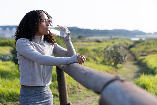 African American woman in grey sportswear leaning on rail at coast trail sipping clear water bottle