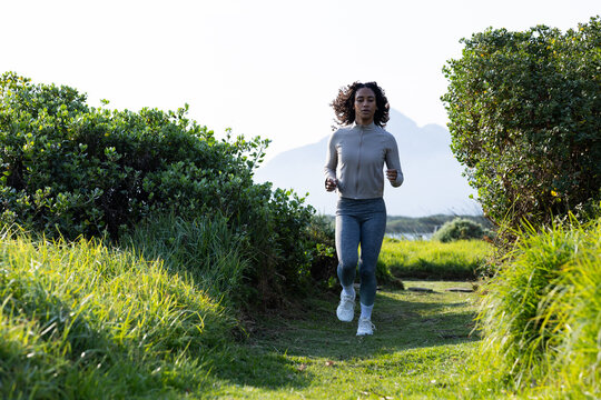 African American woman jogging on grassy trail at coastal reserve in gray jacket and white shoes