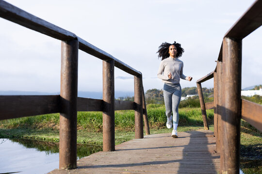 African American woman running on wooden footbridge at park wearing gray jacket
