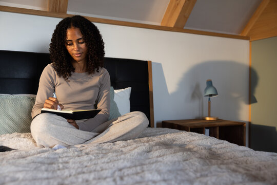 Adult African American woman in beige top sitting cross-legged on bed writing in black notebook