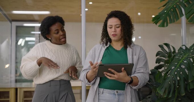 Female coworkers walking in office by plant, woman coming, speaking, tablet tapping to align work