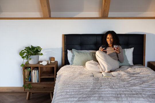 African American woman sitting on bed at home in light gray loungewear holding smartphone and glass