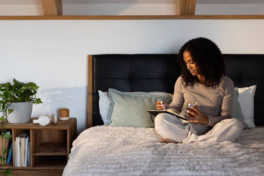 African American woman sitting on bed holding glass mug and journal in bedroom, copy space