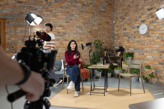 Diverse coworkers filming podcast in studio, presenter in burgundy top holding smartphone, camera