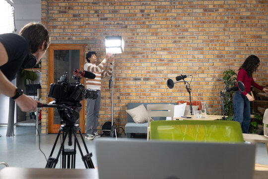 Diverse crew adjusting LED panel, operating camera on tripod, arranging laptop and props in lounge