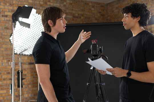 Two men wearing black tops gesturing and holding script in small studio with camera tripod