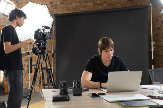 Diverse male coworkers operating camera and working on laptop in studio with softbox, lenses