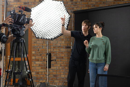 Male director gesturing, woman in green knit posing in studio beside camera and softbox, copy space
